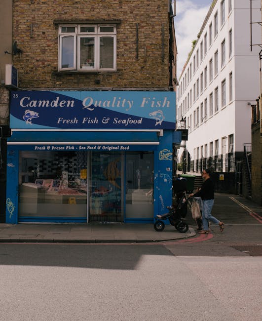 A street-level view showing the front of a fish and seafood shop named Camden Quality Fish, with a blue storefront sign and large glass display window featuring packaged seafood products. Above the sh